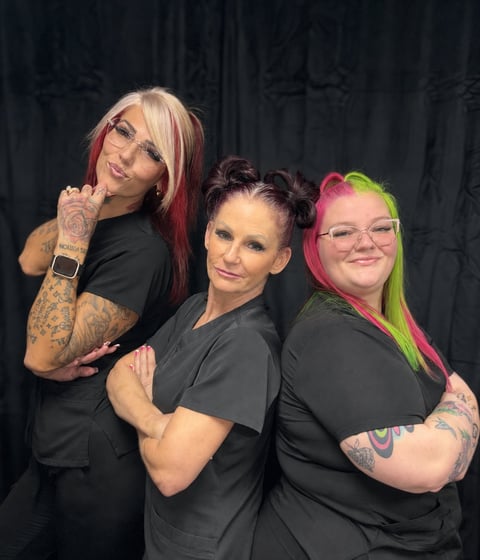 Three women wearing black shirts posing together against a dark background. All have tattoos and colorful hair styling in pink, red, and green tones.