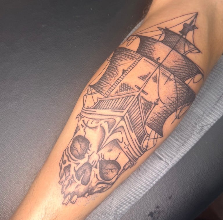 Black ink tattoo on forearm featuring a sailing ship above a decorative skull design