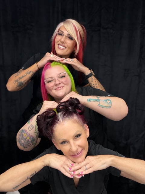 Three smiling women with colorful hair and tattoos posing together against a dark background