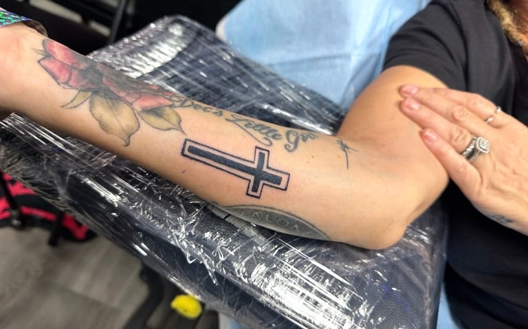 Close-up of person's arm showing a cross tattoo and a floral tattoo design, wearing a ring and blue shirt