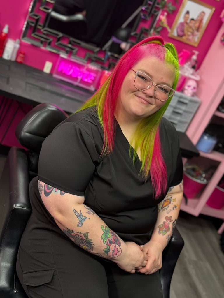 Sara Lee, custom tattoo artist at CC Styling Studio in Bradenton, FL — smiling with pink and yellow hair, glasses and a black shirt in a neon pink-lit room