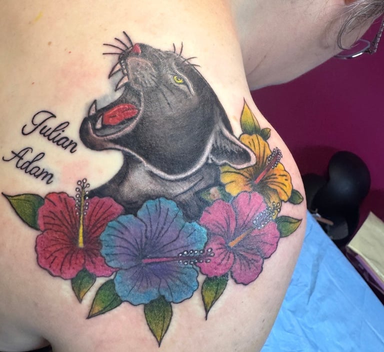 Tattoo of a black panther with red mouth surrounded by colorful flowers and butterfly on a person's shoulder