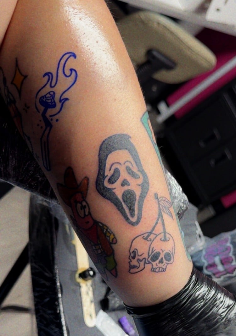 Arm with multiple horror-themed tattoos including a ghost face from Scream, a blue tribal design, and a skull with flowers