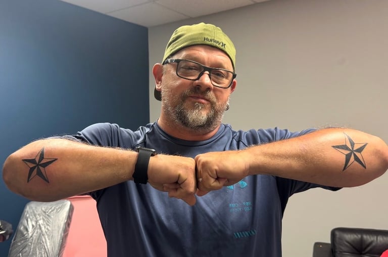 Man with glasses and yellow cap posing with arms outstretched showing star tattoos on both forearms