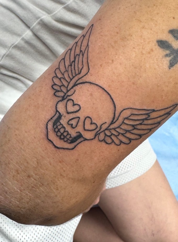 Black outline tattoo of a winged skull with heart eyes on someone's upper arm