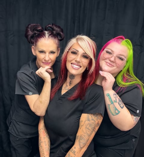 Three smiling women with colorful hair and tattoos posing together against a dark background