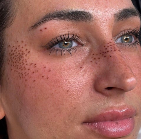 Close-up of a persons face showing freckles, defined eyebrows, and brown eyes with makeup