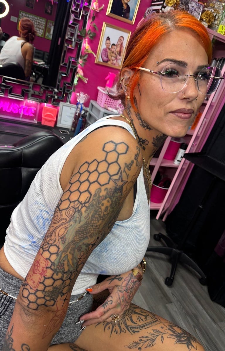 Woman with orange hair and glasses displaying arm tattoos in a pink neon-lit room