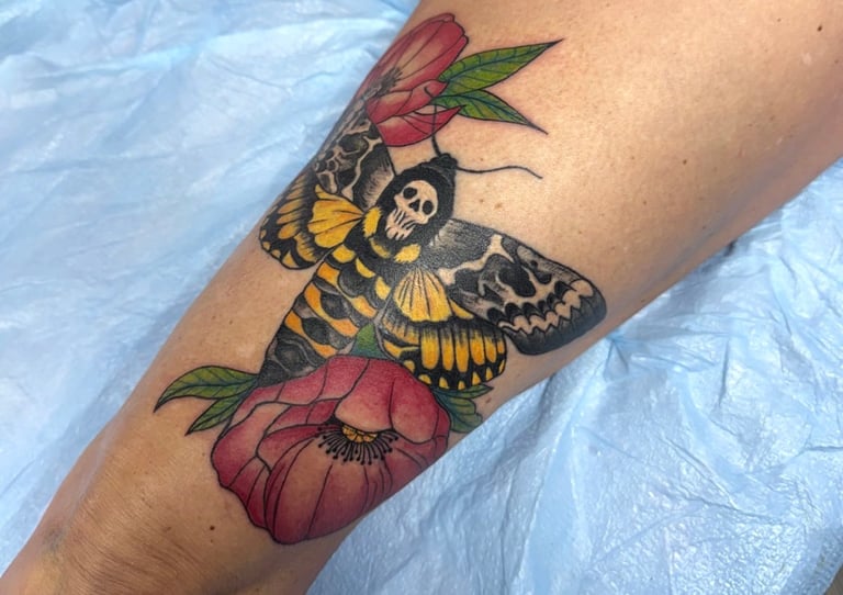 Colorful tattoo on forearm featuring a yellow and black moth with red and pink flowers on blue fabric background