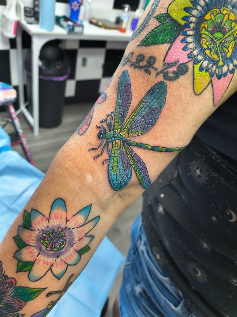 Colorful arm tattoo featuring dragonfly, sunflower, and decorative insects on skin in tattoo parlor setting