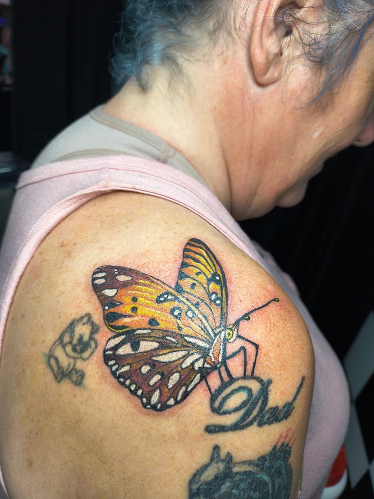 Close-up of a colorful butterfly tattoo on a person's shoulder, featuring yellow and black wings with decorative script lettering below