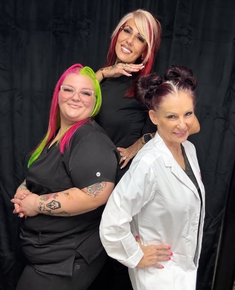 Three women posing together against a dark background. Left woman has pink and lime green hair and wears black. Middle woman has pink and blonde hair in black. Right woman wears white blazer with dark updo hairstyle.
