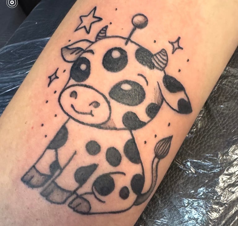 Black ink tattoo of a cute cartoon cow with spots, antennae, stars, and moon elements on a person's arm