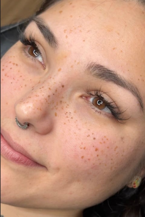 Close-up portrait of a womans face with freckles, defined eyebrows, and a septum piercing, looking downward