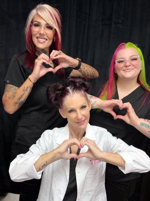 Three women making heart shapes with their hands, with colorful hair including pink and green, posing together against a dark background