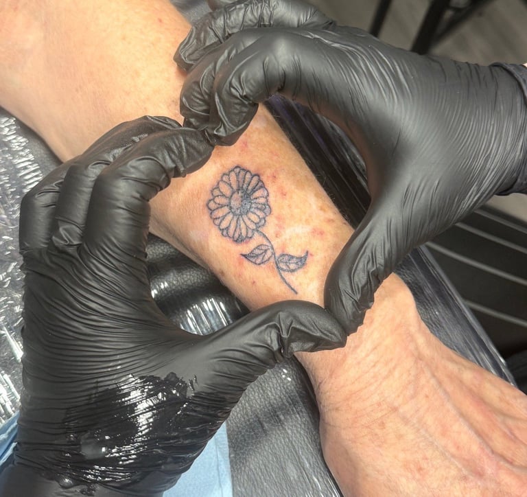 Tattoo artist wearing black gloves working on a small blue flower tattoo on a person's forearm