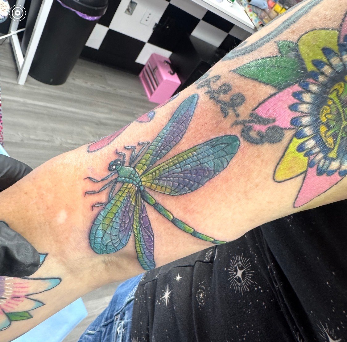 Colorful dragonfly tattoo on forearm with purple, blue, green, and yellow wings in a tattoo studio