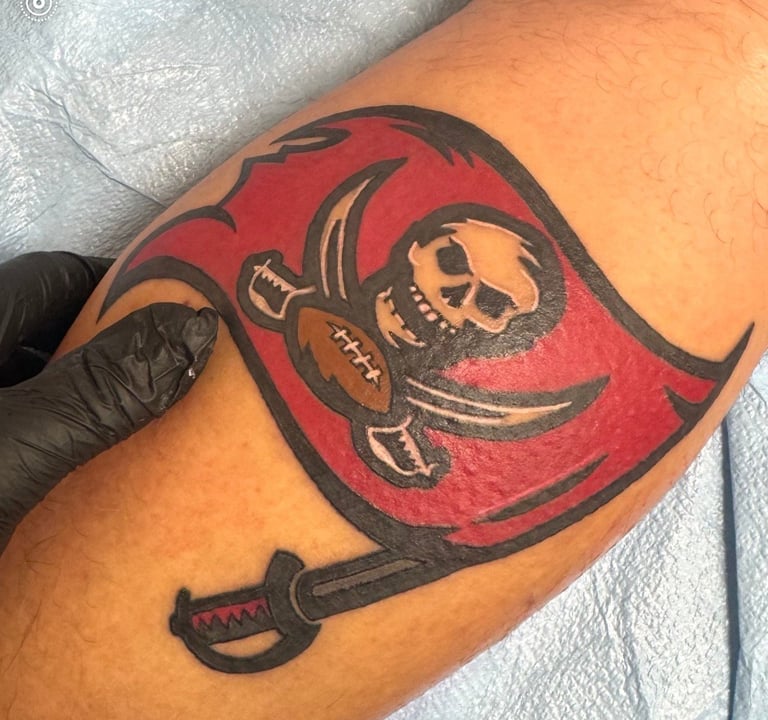 Fresh red and gold skull pirate flag tattoo on forearm with black outlines and artist wearing glove during application