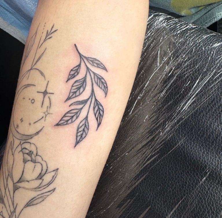 Arm tattoo featuring a crescent moon face with leaves and botanical elements in black ink
