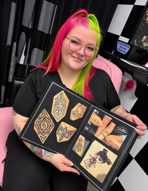 Person with hot pink and lime green split hair wearing glasses and black shirt displays a tattoo portfolio book with various ornamental and decorative designs