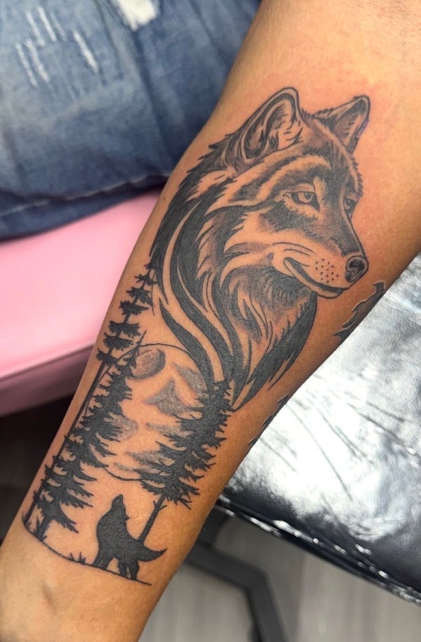Black ink tattoo on forearm featuring a wolf head above an evergreen forest and silhouetted hiking figure
