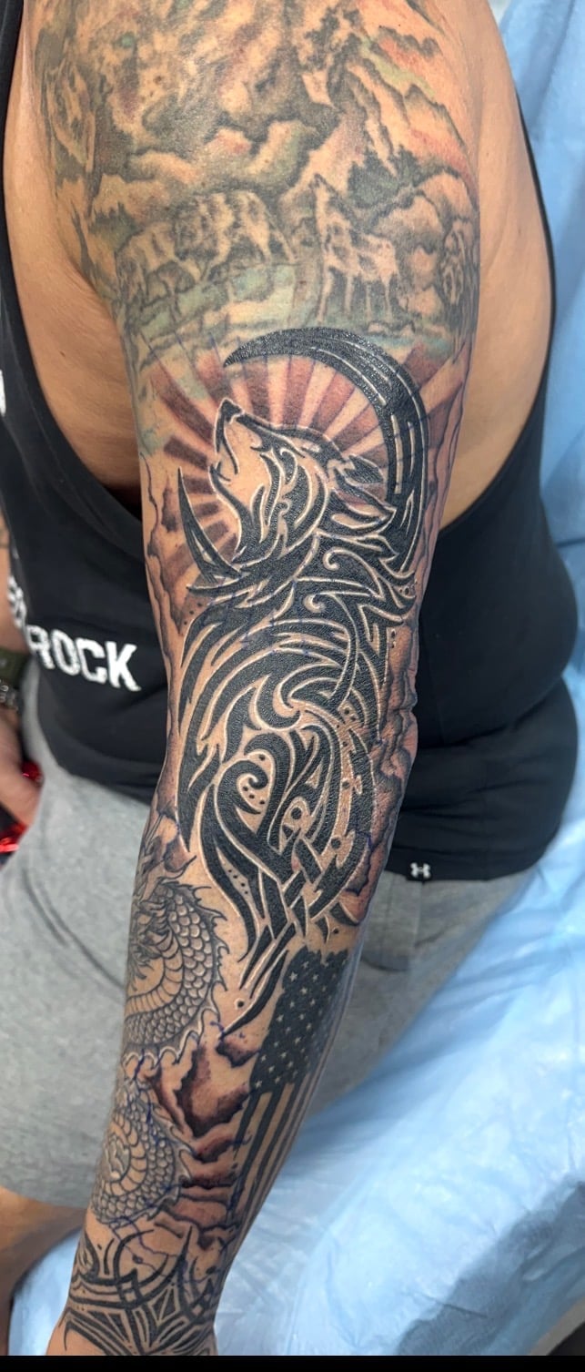 Full sleeve tattoo featuring intricate tribal patterns, a wolf head with sun rays, dragon scales, and ornamental designs in black ink on a person's arm