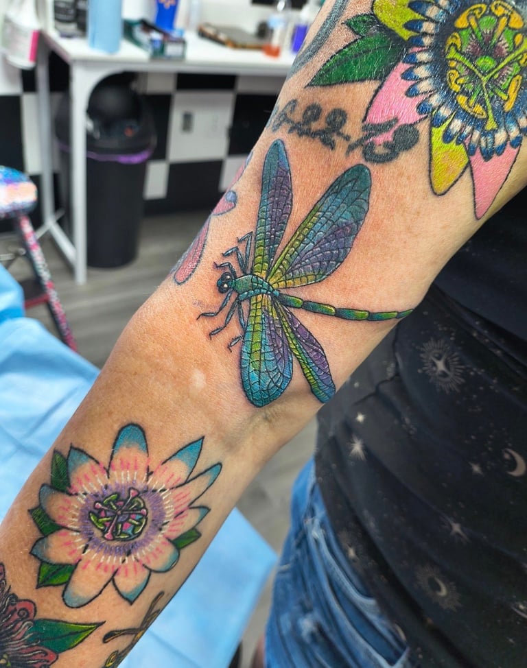 Colorful tattoo sleeve on arm featuring green and blue dragonfly, pink and yellow flowers, and nature-inspired designs