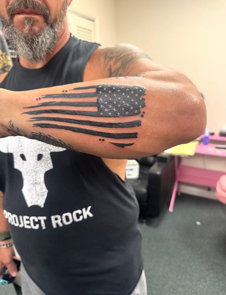 Man displaying an American flag tattoo on his forearm with horizontal stripes and stars, wearing a Project Rock shirt