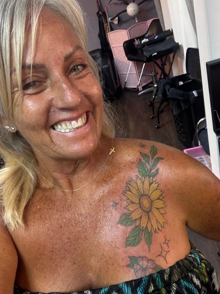 Woman with blonde hair smiling at camera, displaying shoulder tattoos of sunflowers and leaves in a studio setting