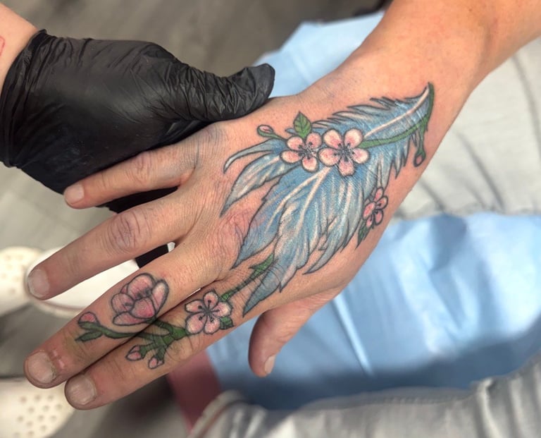 Tattoo artist wearing black gloves examines a colorful floral hand tattoo on a client's hand during a tattoo session