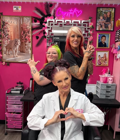 Three women posing in a vibrant pink salon setting, with neon heart sign and beauty decor on walls