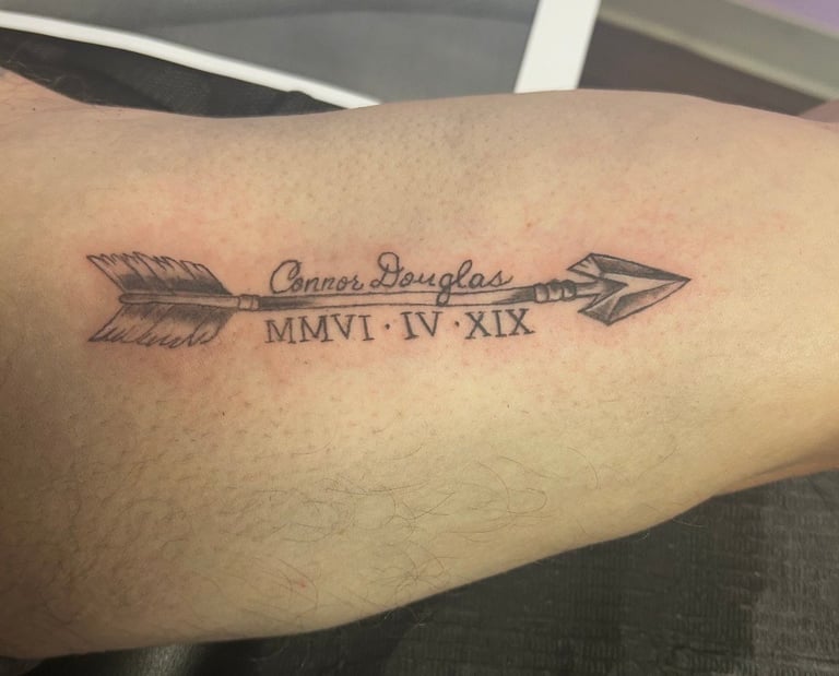 Arrow tattoo on forearm with Connor Douglas name and Roman numerals MMVI IV XIX