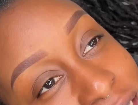 Close-up of a womans face with professionally shaped eyebrows, defined eye makeup, and a nose piercing, looking toward camera